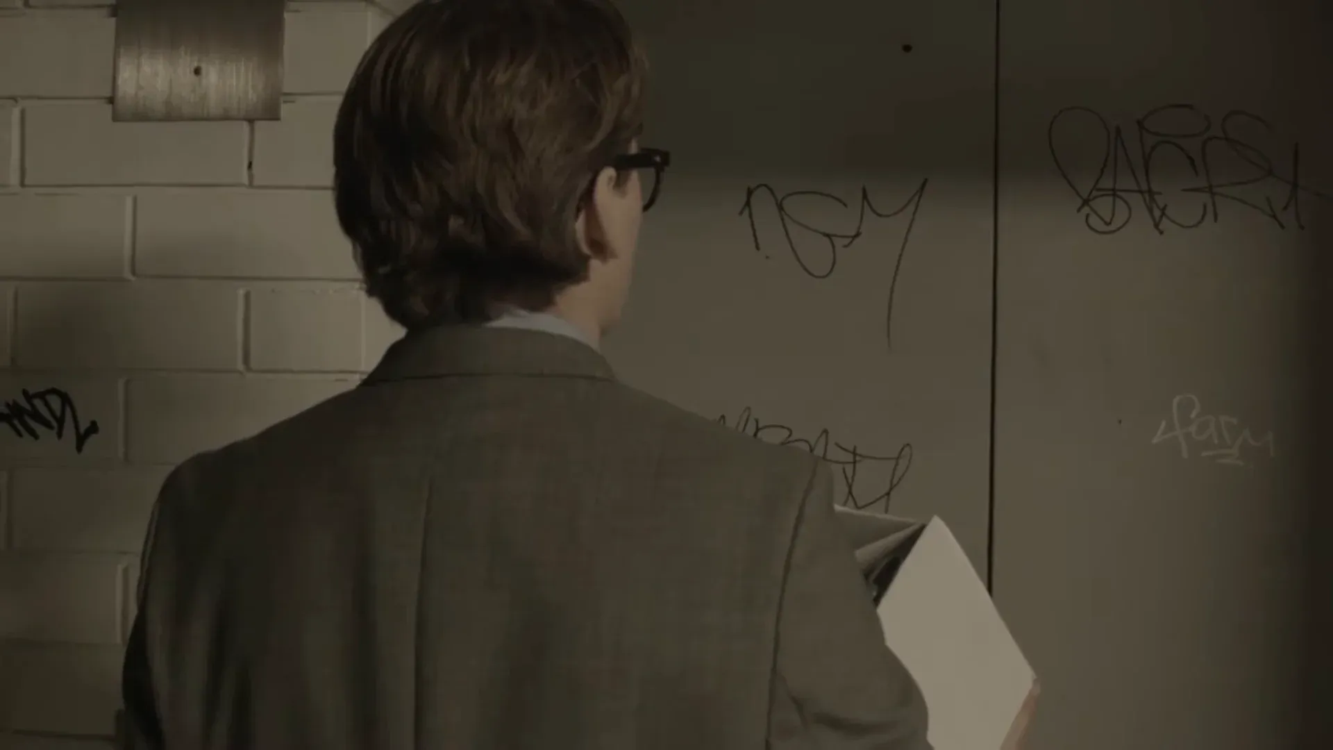 A man in a grey suit, seen from behind, facing a tagged concrete wall in a stairwell, holding a blank sheet of paper at his side.