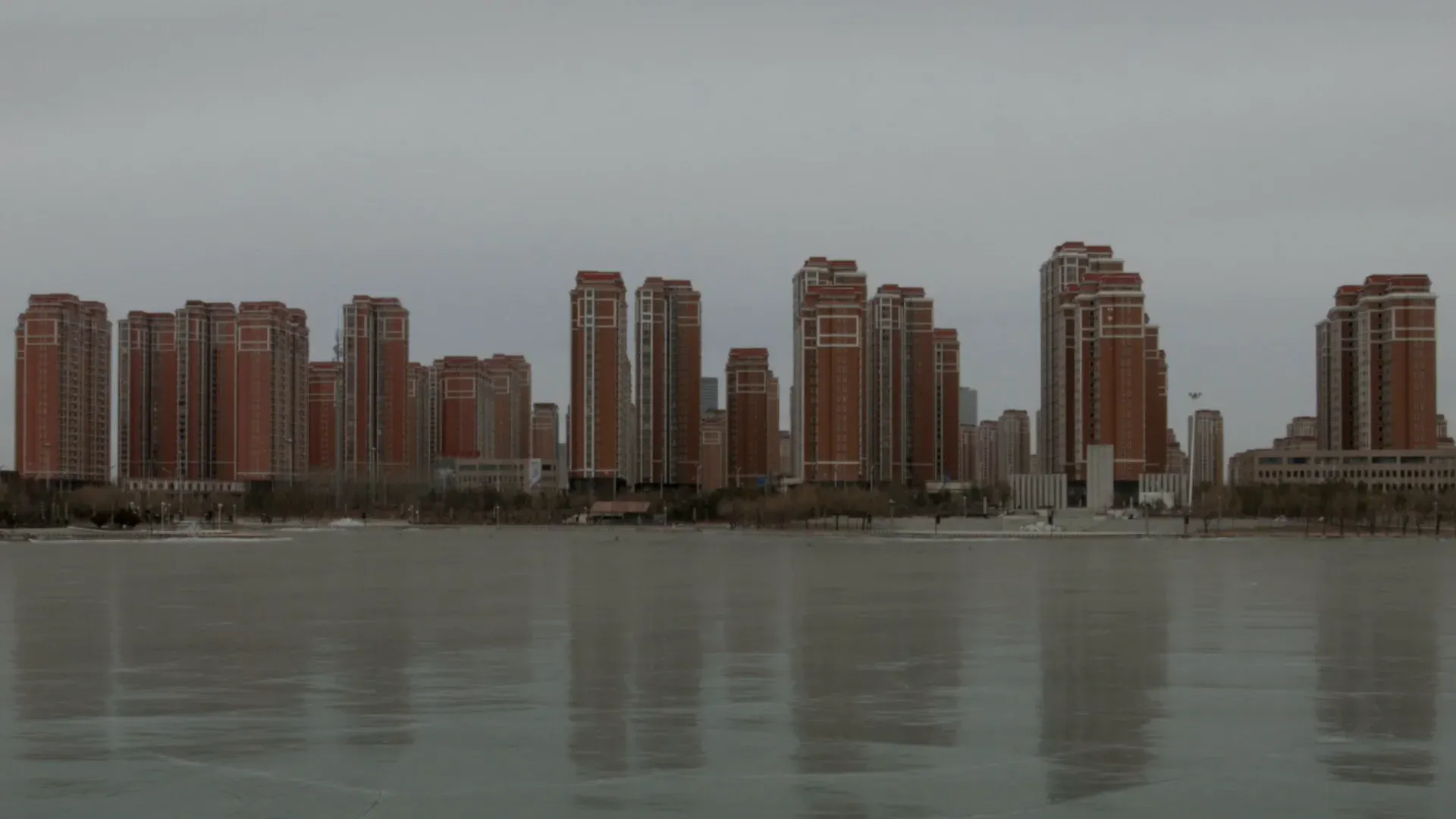 A row of identical high-rise towers across flat water under a hazed sky, no one in sight.