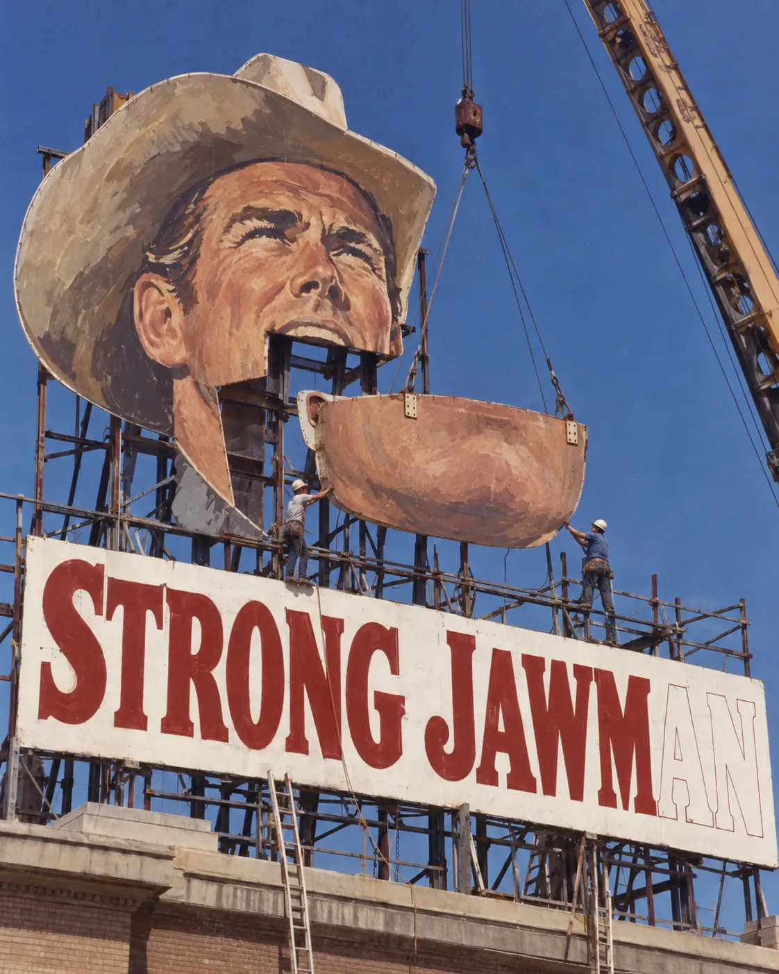 A photograph that looks like 1970s slide film: riggers craning an enormous painted jaw into place on a rooftop billboard of a cowboy, above the words STRONG JAWMAN with the last two letters unpainted. The image is machine-generated; the billboard never existed.